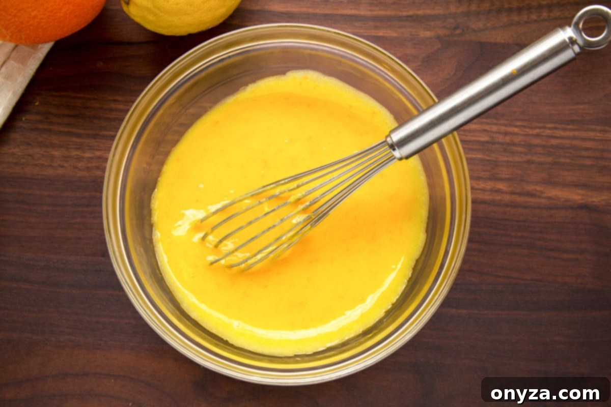 Smooth, golden orange curd in a glass bowl with a stainless steel whisk resting inside, set on a dark wooden cutting board, showcasing the finished curd.