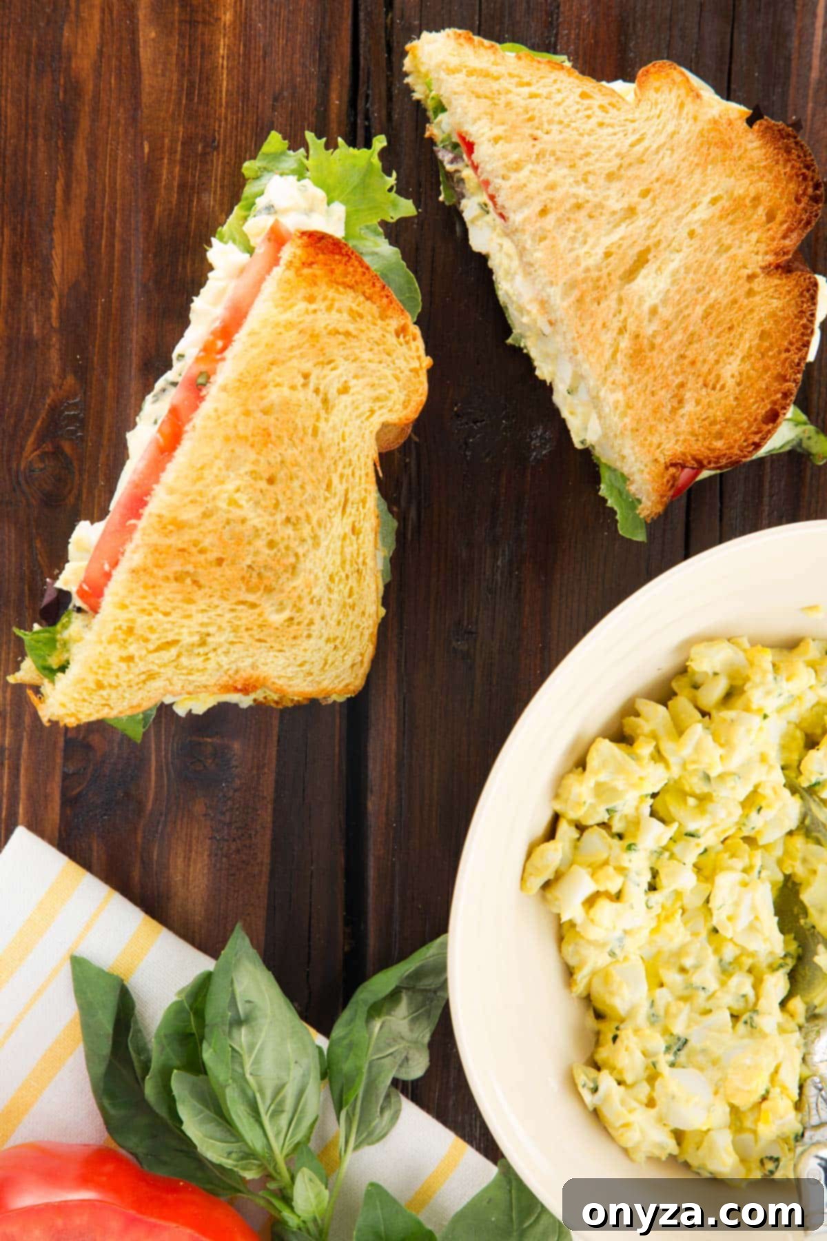 Two halves of a basil egg salad sandwich on brioche toast on a wood board next to a bowl of egg salad, ready to be enjoyed.