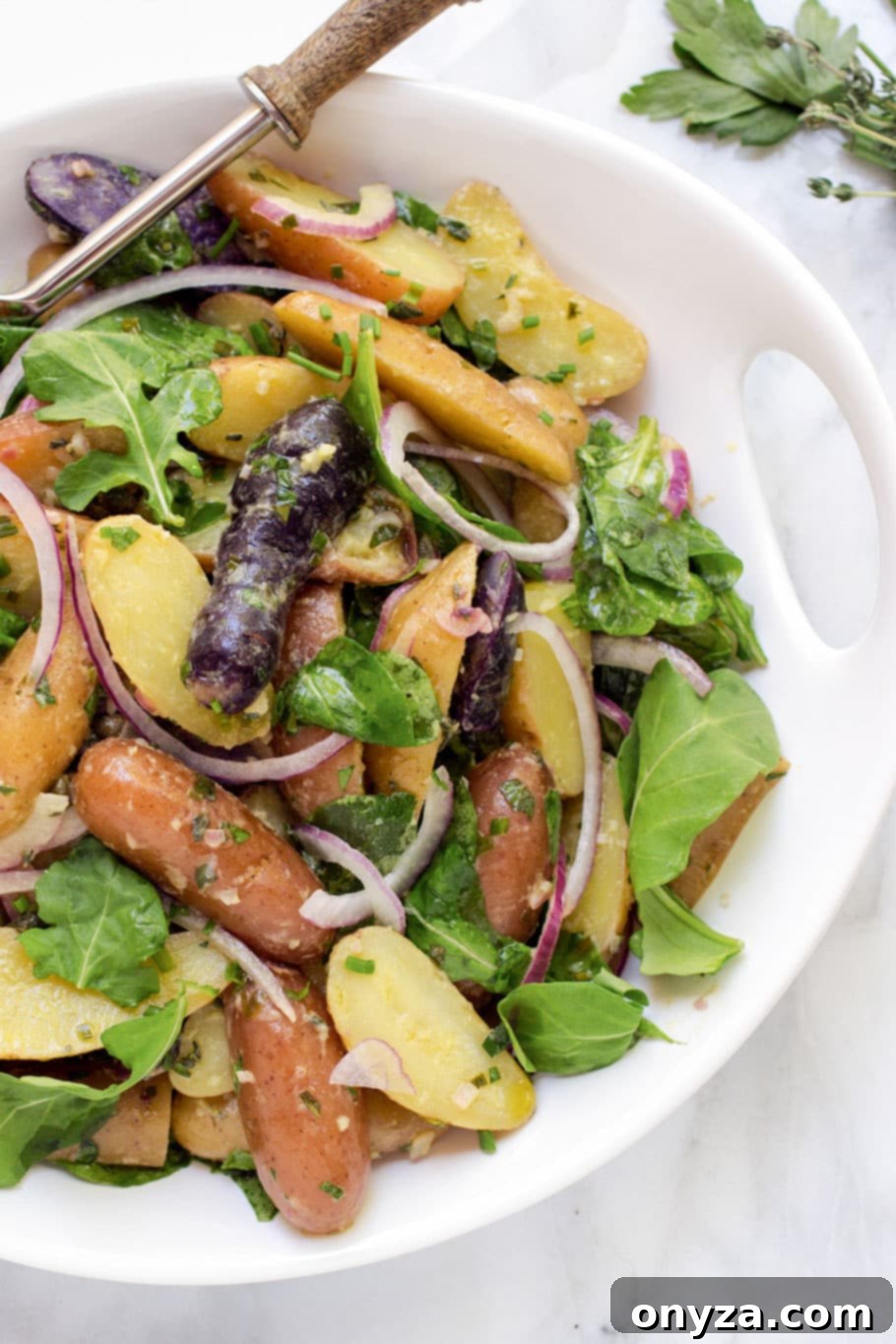 Fingerling potato salad with red onions and baby arugula in a white bowl with a serving spoon, garnished with fresh herbs.
