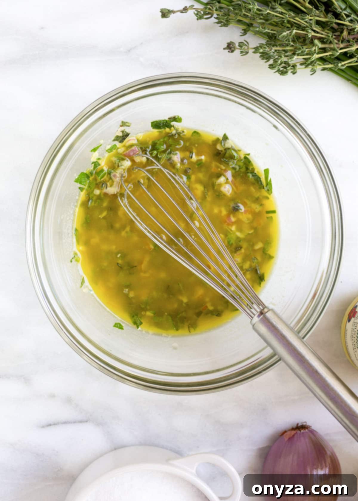A glass bowl filled with a vibrant herb vinaigrette, with a small stainless steel whisk resting inside, on a white marble board.