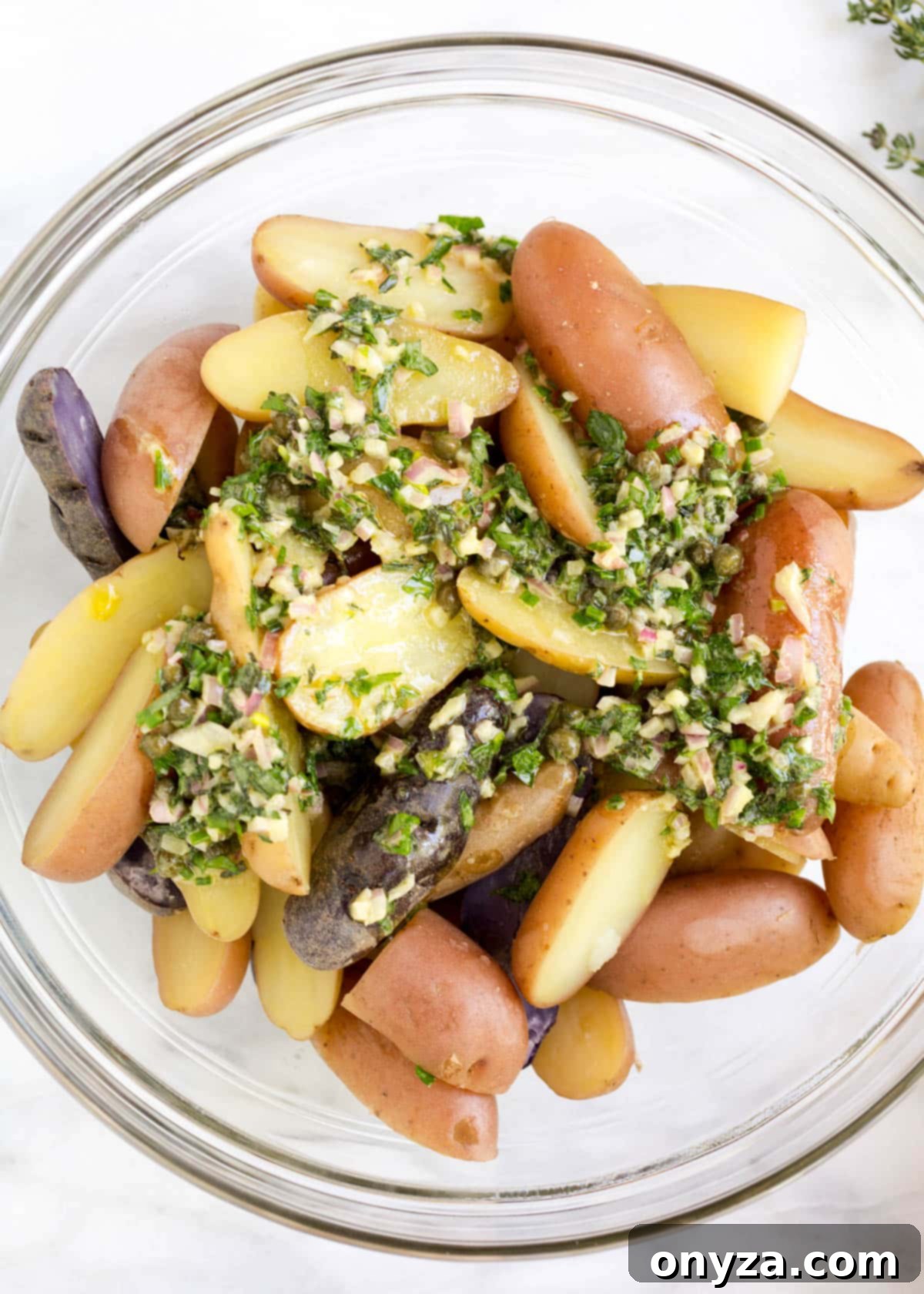 Warm, cooked fingerling potatoes topped with a generous amount of herb vinaigrette in a clear glass bowl, resting on a white marble board.