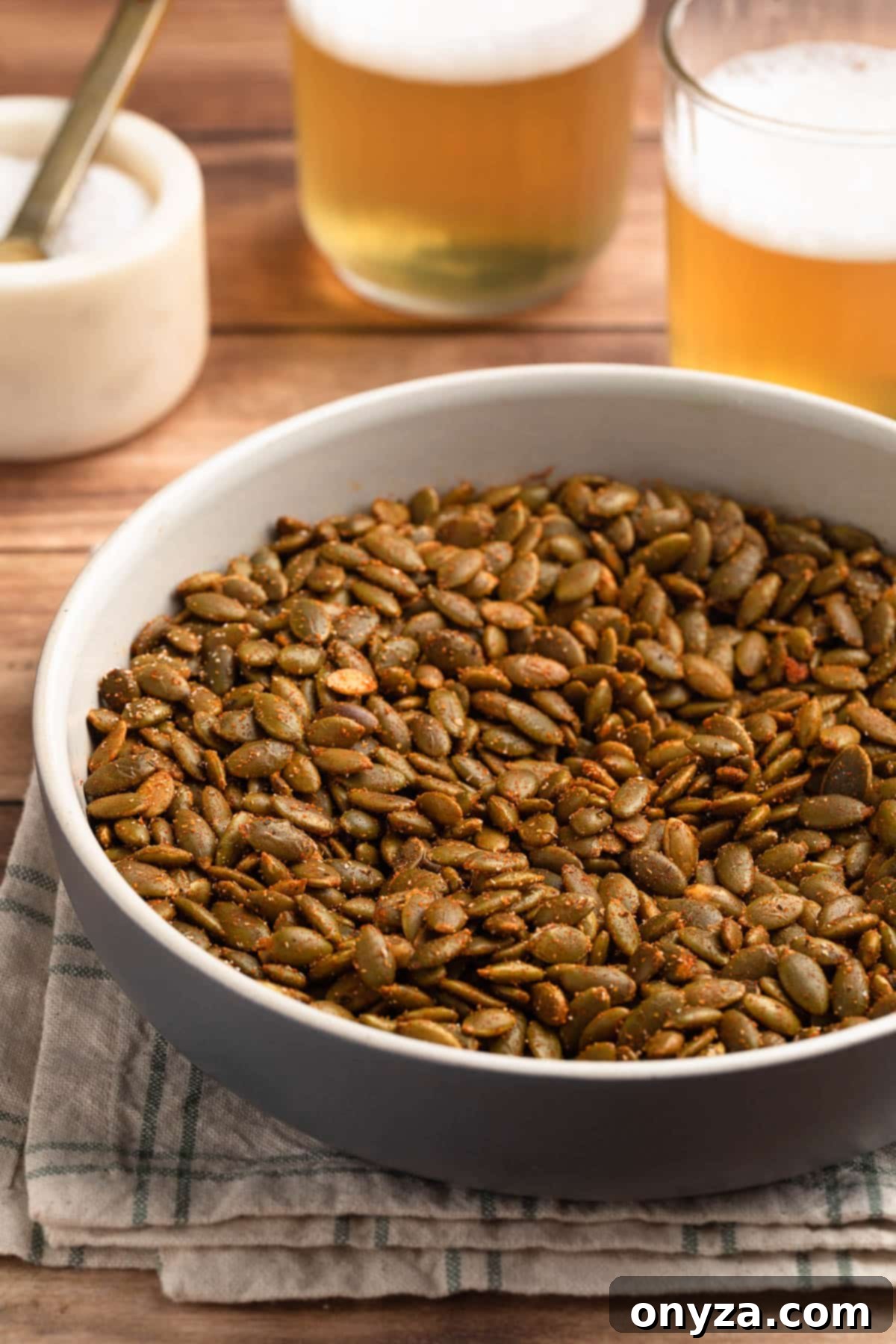 A ceramic bowl filled with golden-brown spiced pepitas, flanked by two small glasses of beer and a marble crock of salt.