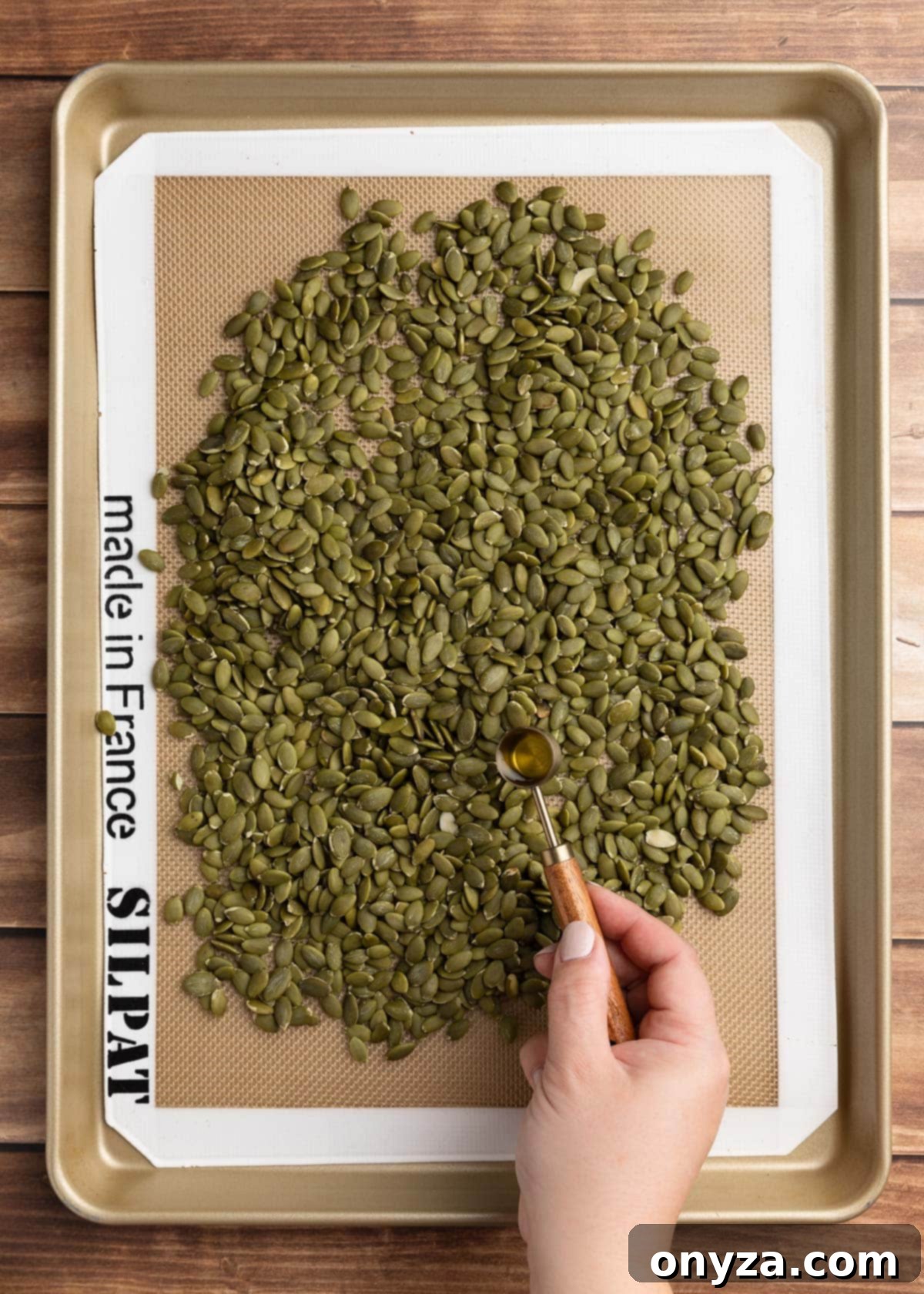 Olive oil being drizzled onto a pan of raw pepitas from a measuring spoon, ensuring an even coating.