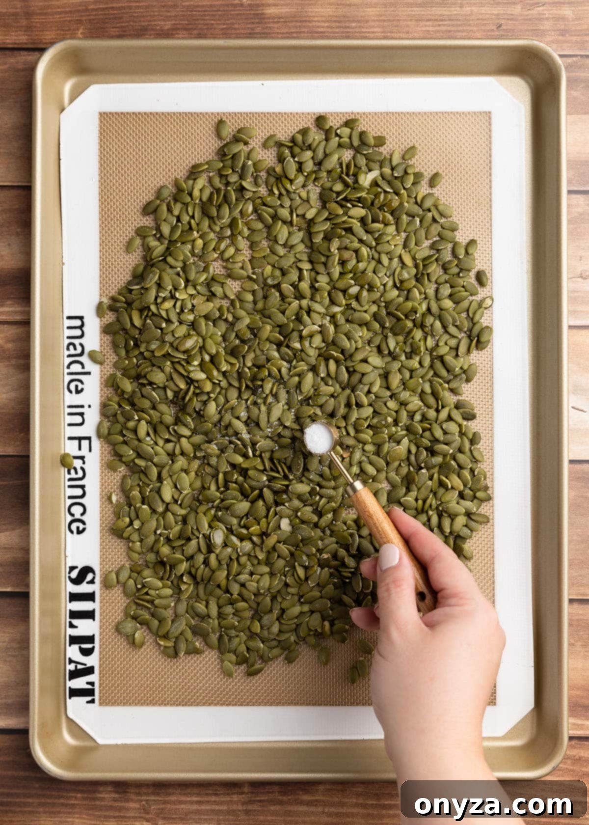 A measuring spoon sprinkling kosher salt onto a pan of raw pepitas, preparing them for roasting.