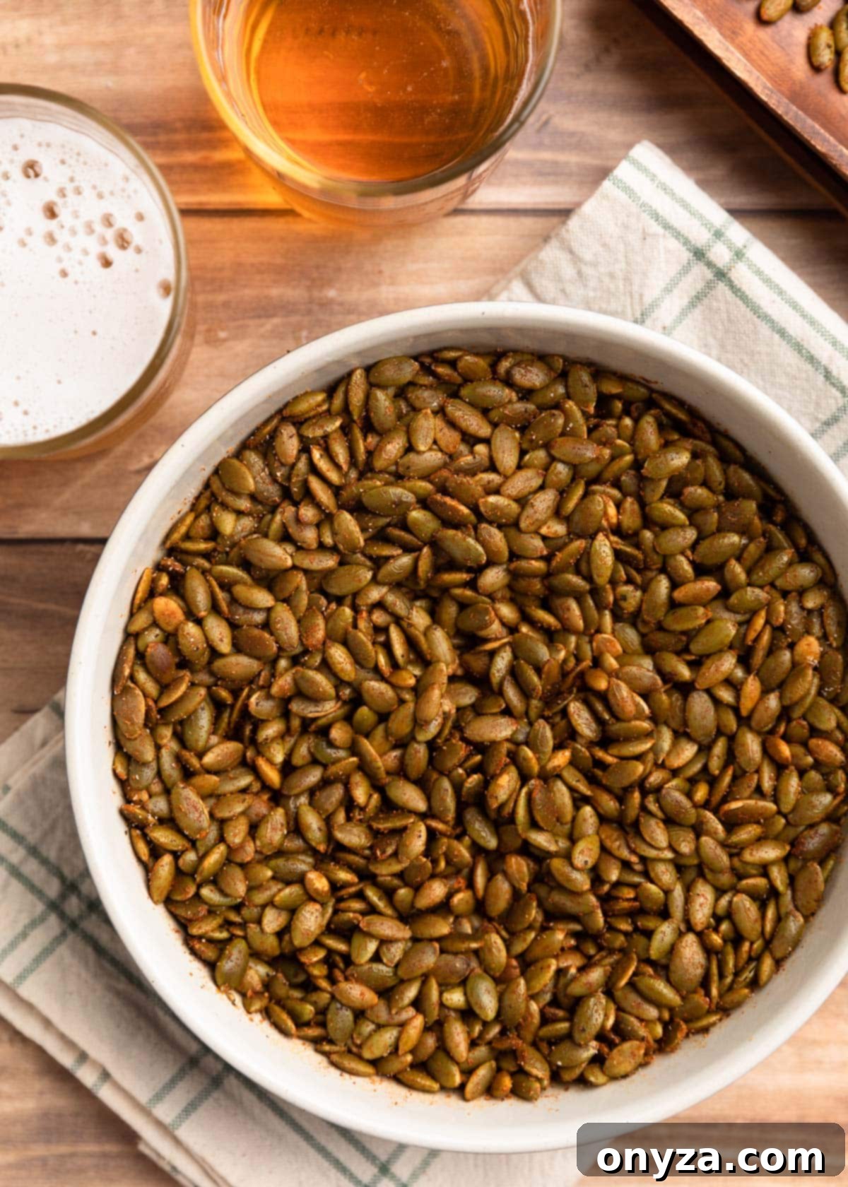 A ceramic bowl brimming with spiced pepitas, elegantly placed beside two glasses of beer and a green and ivory windowpane print napkin.
