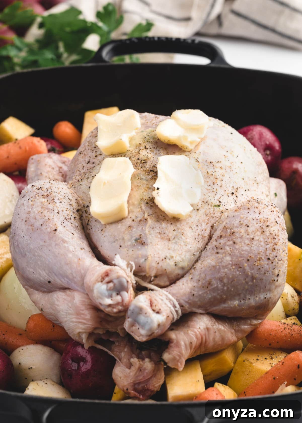 raw whole chicken topped with pats of butter on a bed of root vegetables in a cast iron roasting pan, ready for the oven