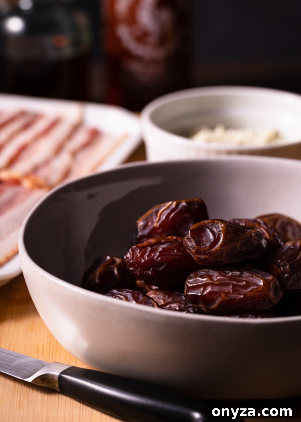 A bowl of fresh Medjool dates is shown alongside a bowl of crumbled blue cheese, with raw bacon strips visible in the background, ready for preparation.