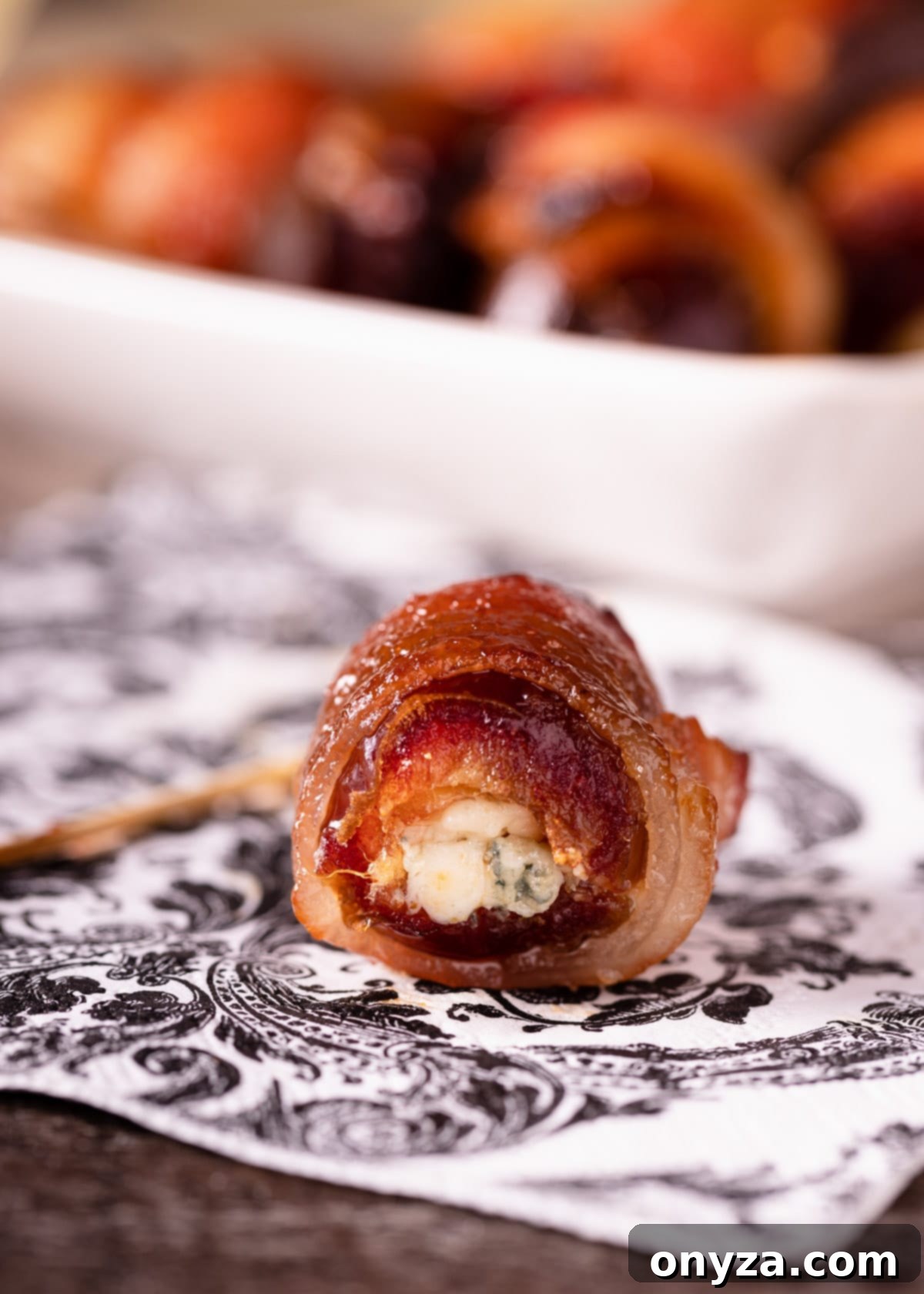 A close-up of a single blue cheese stuffed date, perfectly wrapped in bacon and glazed, resting on a stylish printed napkin.
