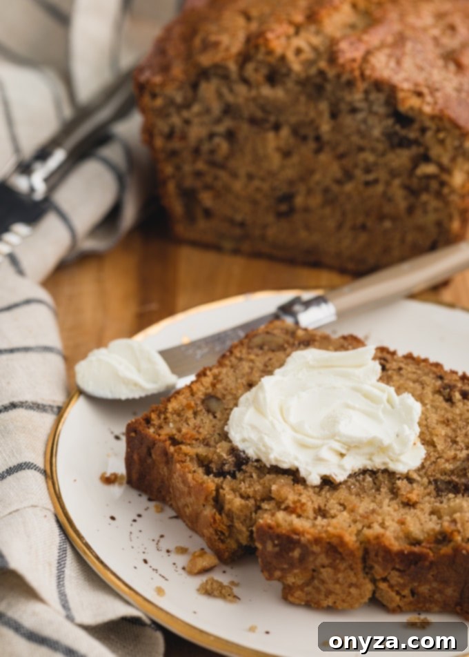 A slice of brown butter banana bread topped with a generous spread of cream cheese on a white plate, ready for a delightful bite.