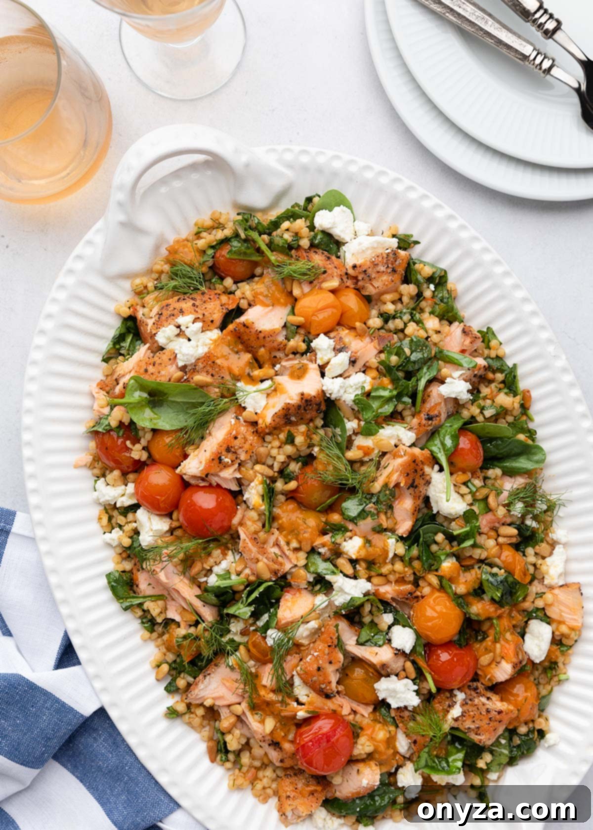 Flaked Salmon Couscous Salad with Sun-Kissed Tomato Vinaigrette 2 Fresh salmon and Israeli couscous salad on a white serving platter, garnished with herbs and a blue and white striped napkin