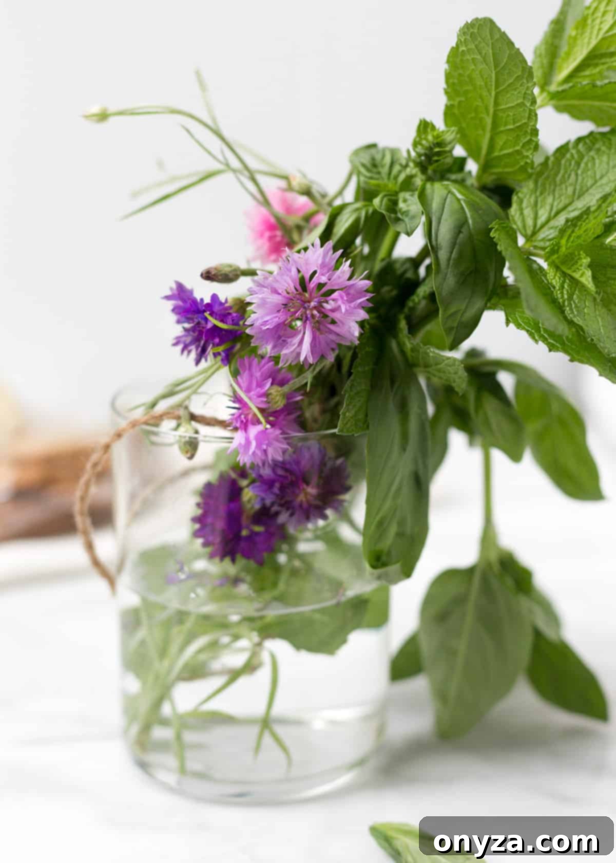 Glass jar filled with fresh basil, mint, and pink and purple edible flowers on a white marble board