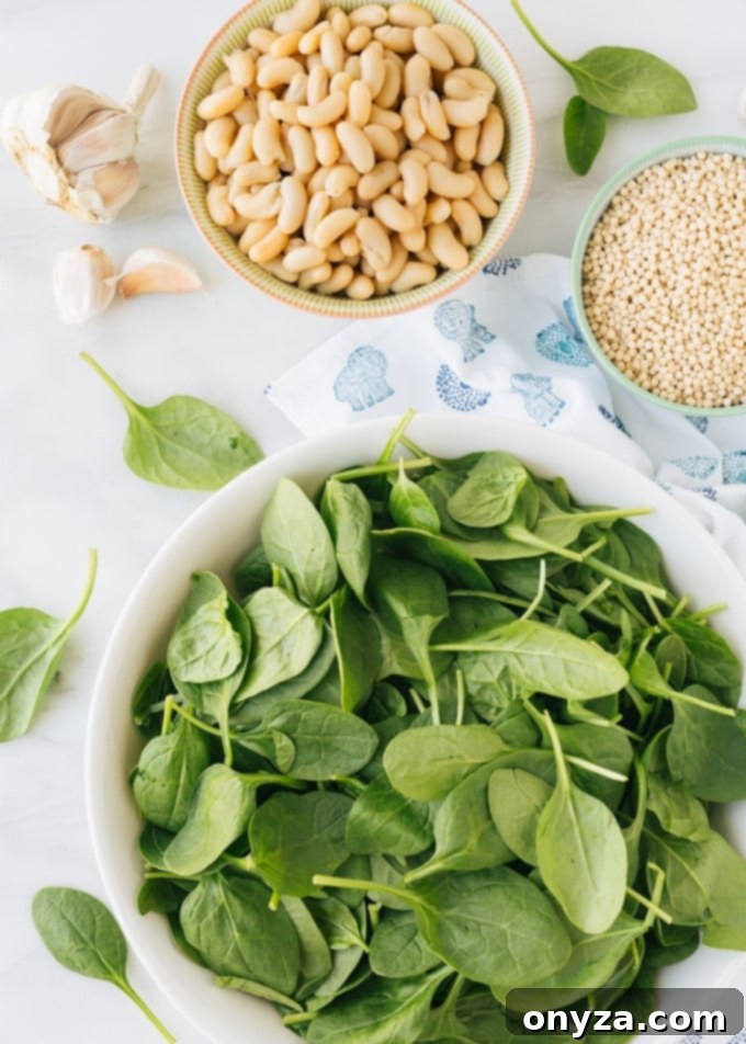 Overhead view of various fresh ingredients for pearled couscous stuffed peppers, including bowls of vibrant baby spinach, tender cannellini beans, and uncooked Israeli couscous pearls.