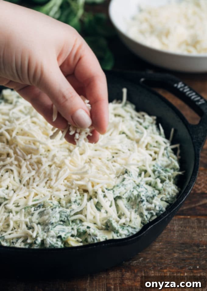 Shredded mozzarella cheese being generously sprinkled over an unbaked six-cheese spinach artichoke dip in a cast iron pan