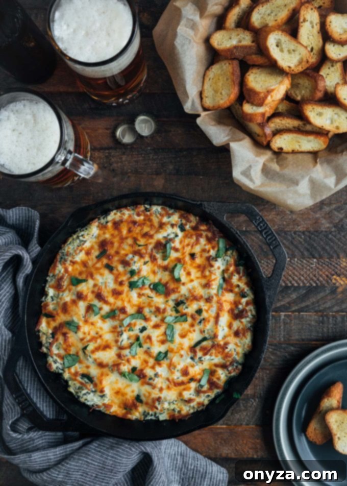 Hot and bubbly six-cheese spinach artichoke dip in a cast iron pan, served with a bowl of crispy crostini