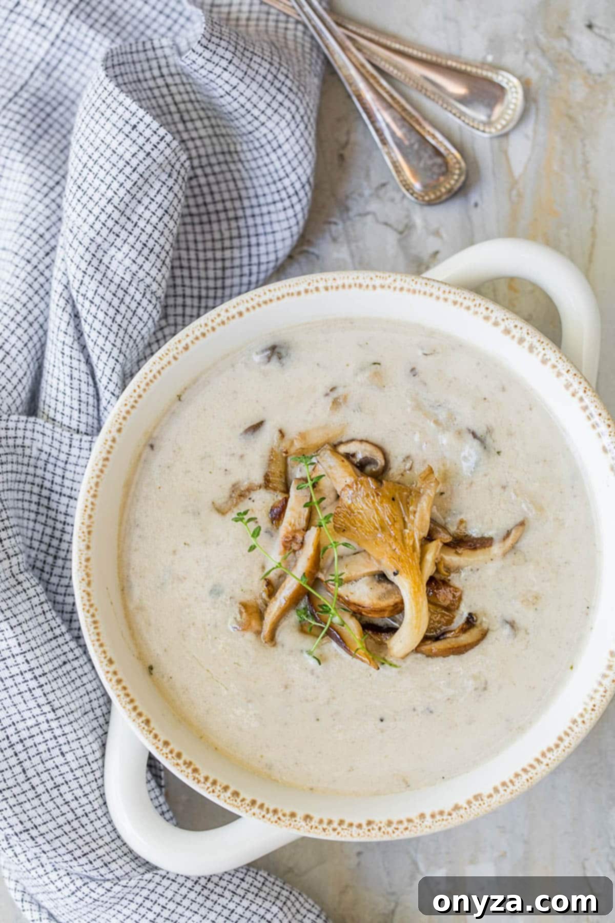 Overhead shot of a single white, double-handled soup bowl filled with elegant cream of mushroom soup, placed next to a blue and white small checked napkin.