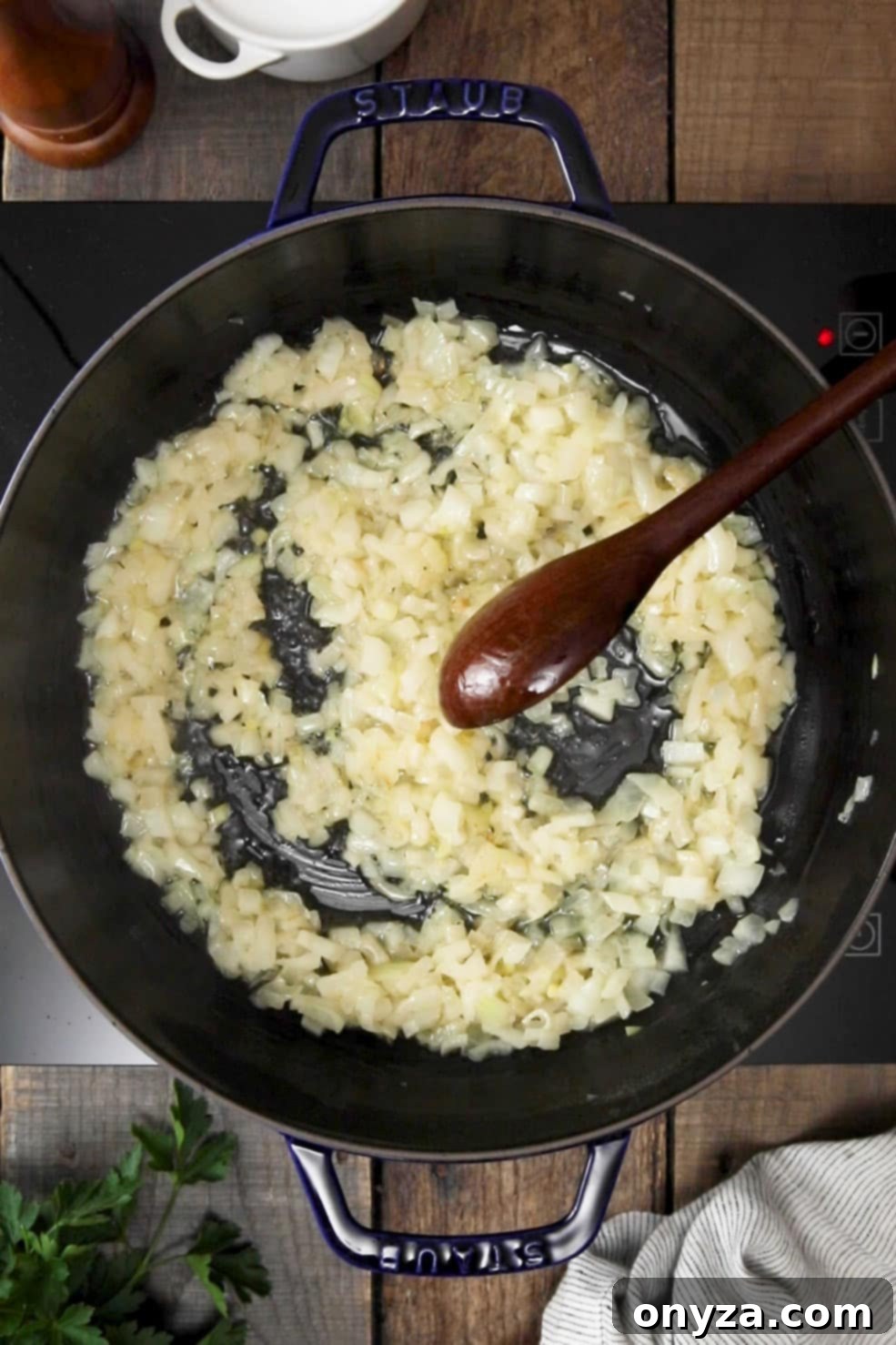 Chopped yellow onions softening in melted butter within a black enameled cast iron Dutch oven, stirred with a wooden spoon.