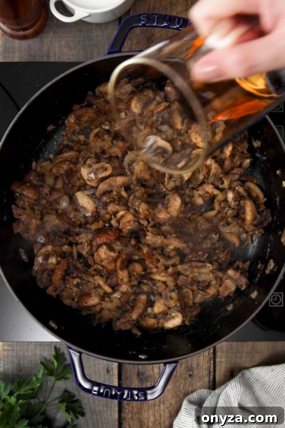 Dry sherry being poured from a glass measuring cup into a black enameled cast iron Dutch oven with cooked mushrooms and onions, initiating deglazing.