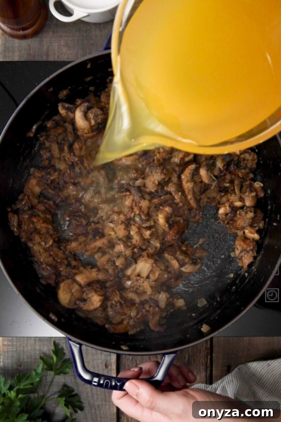 Chicken stock being carefully poured from a liquid measuring cup into sautéed mushrooms and onions in a black enameled cast iron Dutch oven.