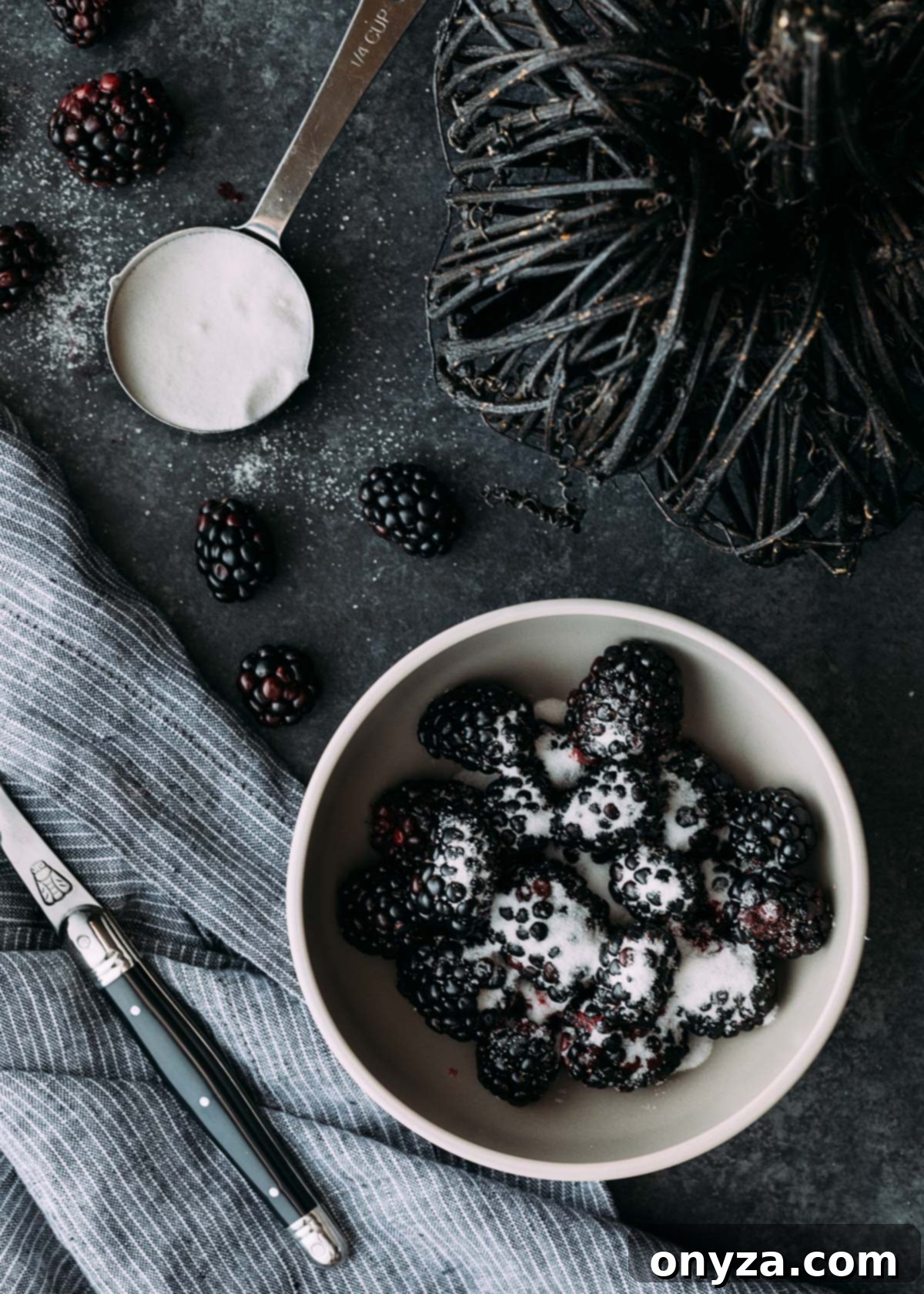 Close-up view of fresh blackberries gently tossed with granulated sugar in a glass bowl, illustrating the maceration process for making homemade blackberry syrup. The sugar crystals glisten on the berries, hinting at the sweet juice being drawn out, a crucial step for the cocktail's rich flavor.