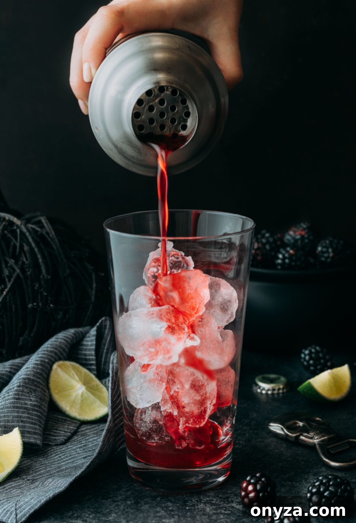 A highball glass filled with ice, ready to receive a freshly shaken Blackberry El Diablo cocktail. A cocktail shaker, still frosty from mixing, is poised above the glass, actively straining the red concoction through a fine-mesh strainer. The backdrop hints at a lively Halloween celebration, emphasizing the drink's festive nature.