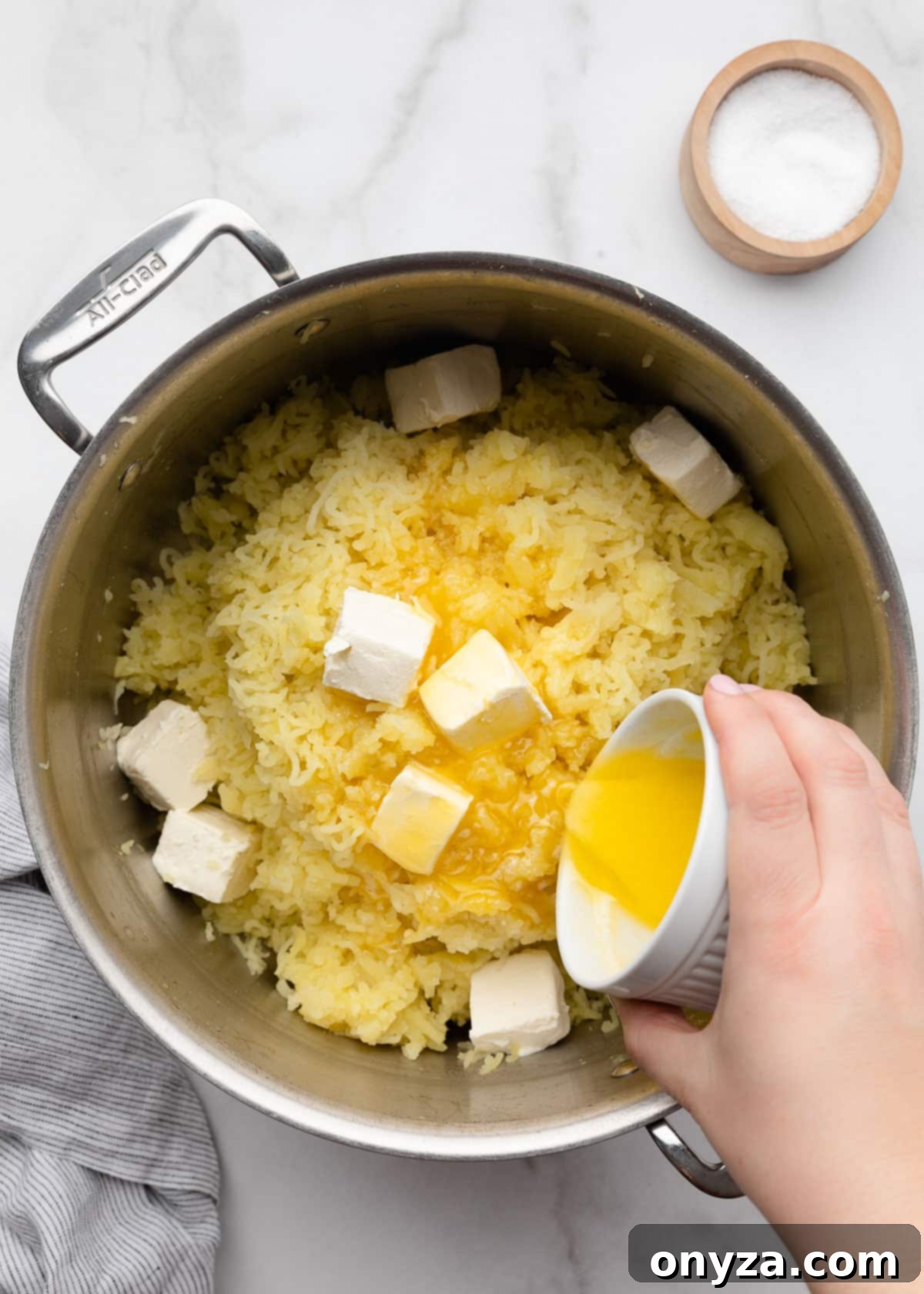 Pouring melted butter into a pot of riced potatoes with cubed cream cheese