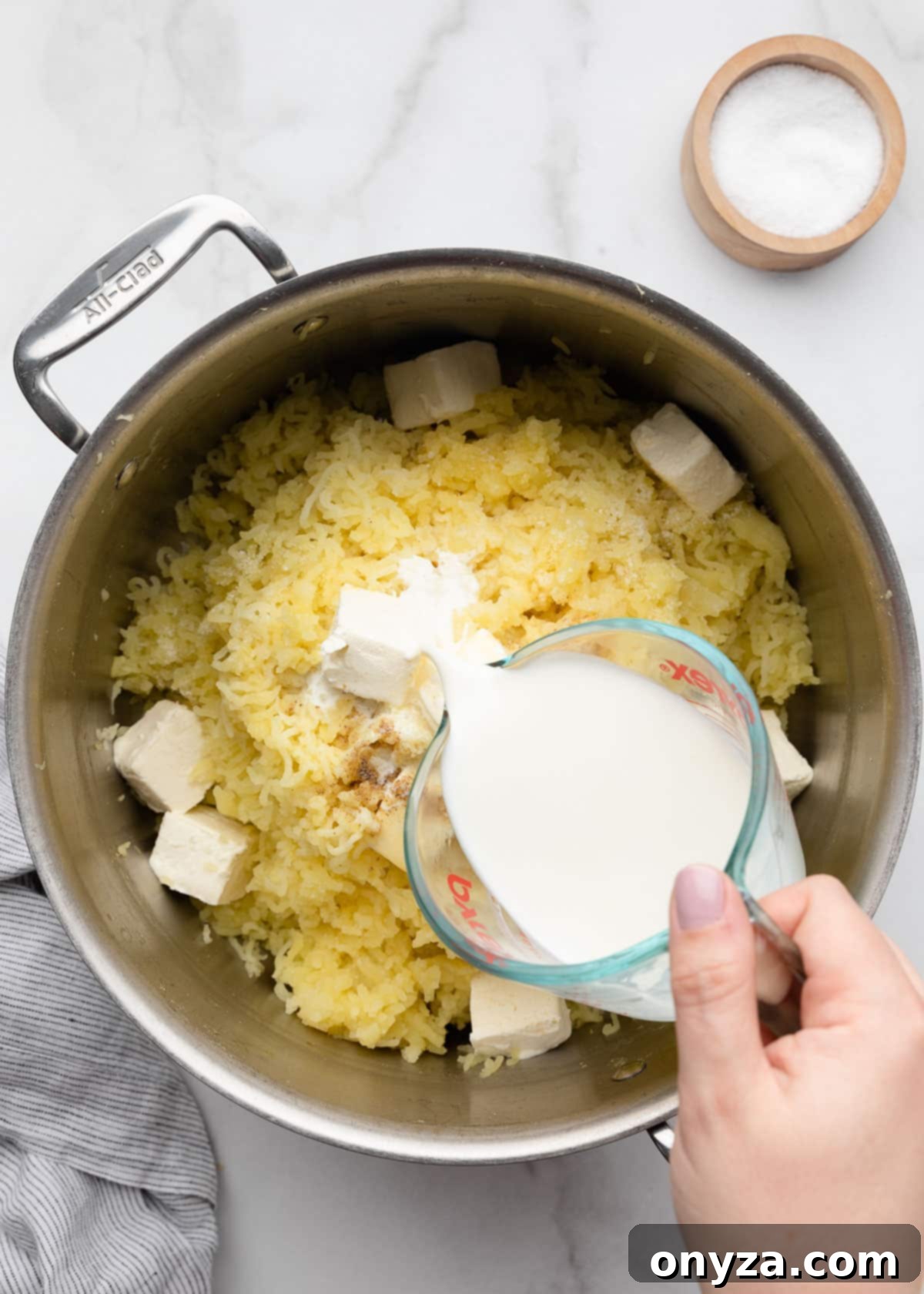 Pouring half and half from a Pyrex measuring cup into a pot of riced potatoes