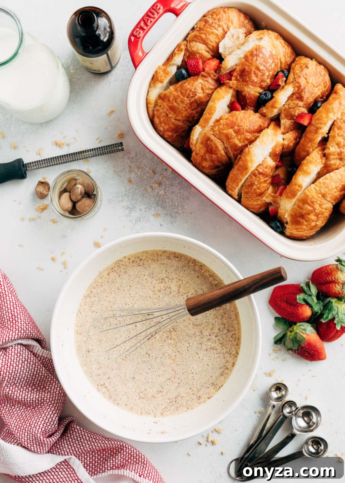 A large bowl containing the freshly whisked French Toast custard mixture, positioned beside a rectangular baking dish already holding neatly sliced croissants and a scattering of fresh berries, all perfectly prepared for the next stage of assembly.