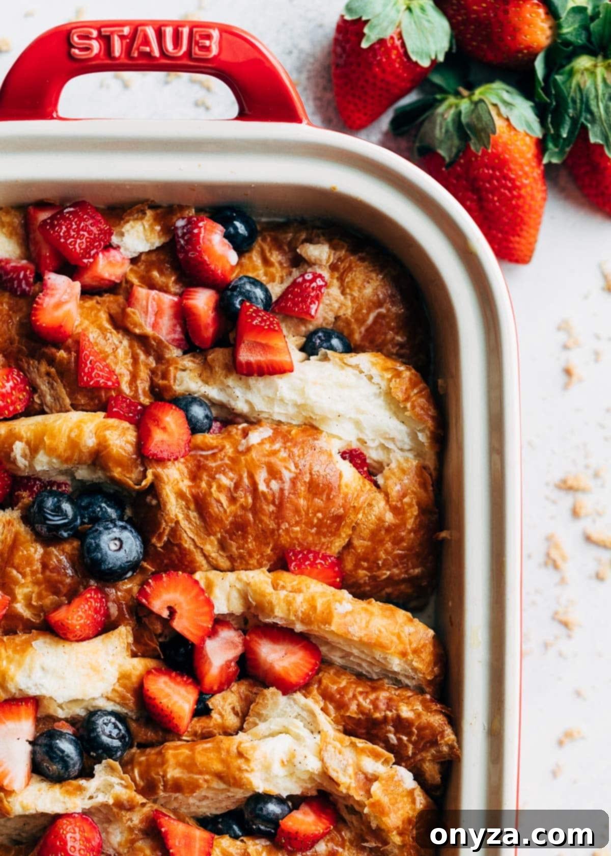 Close-up overhead photo of an unbaked croissant French toast casserole, meticulously arranged with soaked croissant halves and vibrant fresh berries nestled within, all elegantly presented in a rectangular baking dish, awaiting refrigeration.