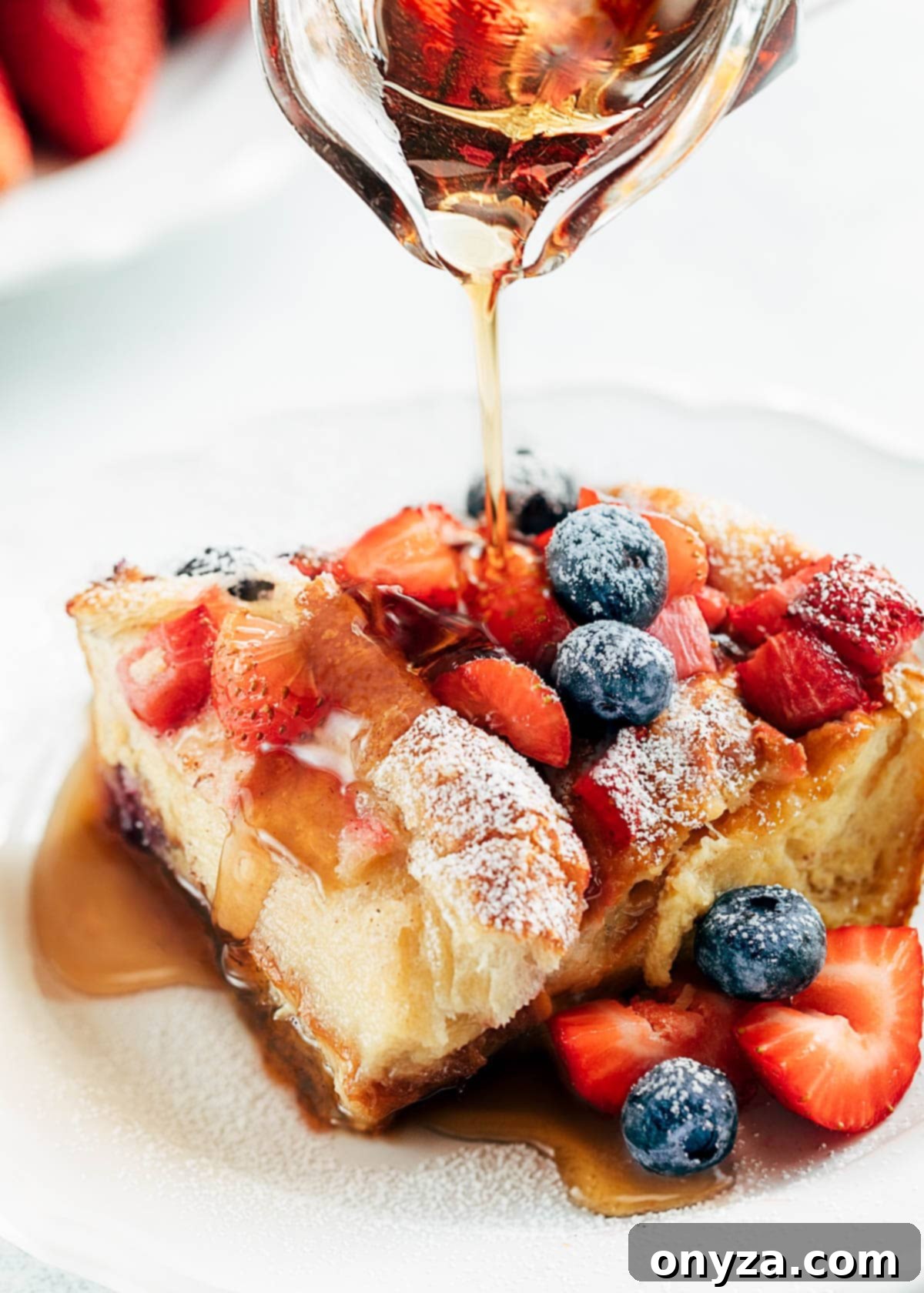 A perfectly portioned wedge of golden-baked croissant French toast casserole topped with a medley of fresh berries and a dollop of whipped cream, beautifully presented on a pristine white plate, with rum-infused maple syrup being artfully drizzled generously over the top.
