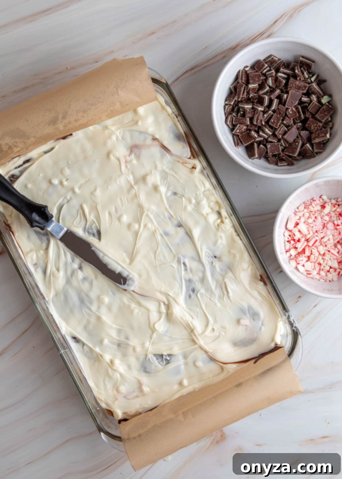 White chocolate spread evenly over the toffee bark, with bowls of chopped Andes mints and crushed peppermint candies nearby.