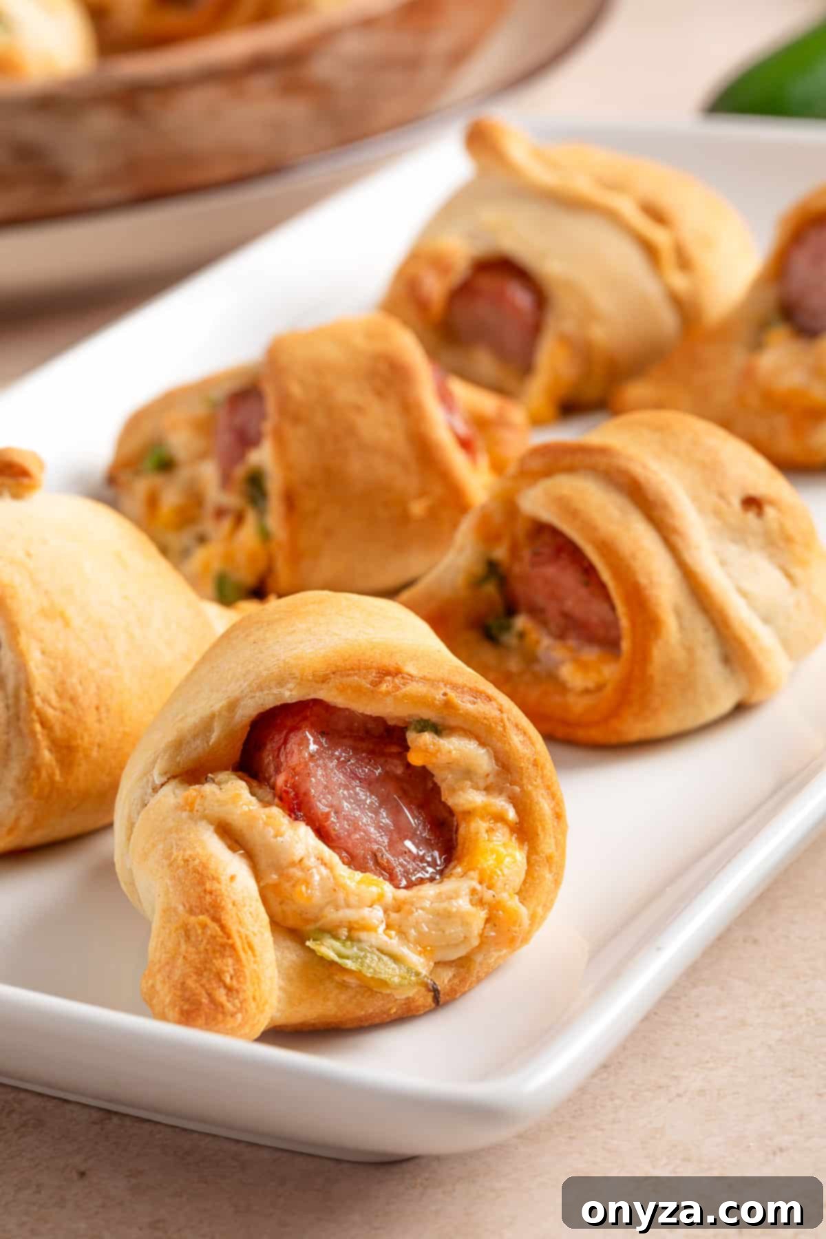 Golden crescent roll-wrapped pigs in a blanket filled with melted cheddar, chopped jalapeño, and smoky cocktail sausages, arranged on a white rectangular serving platter.