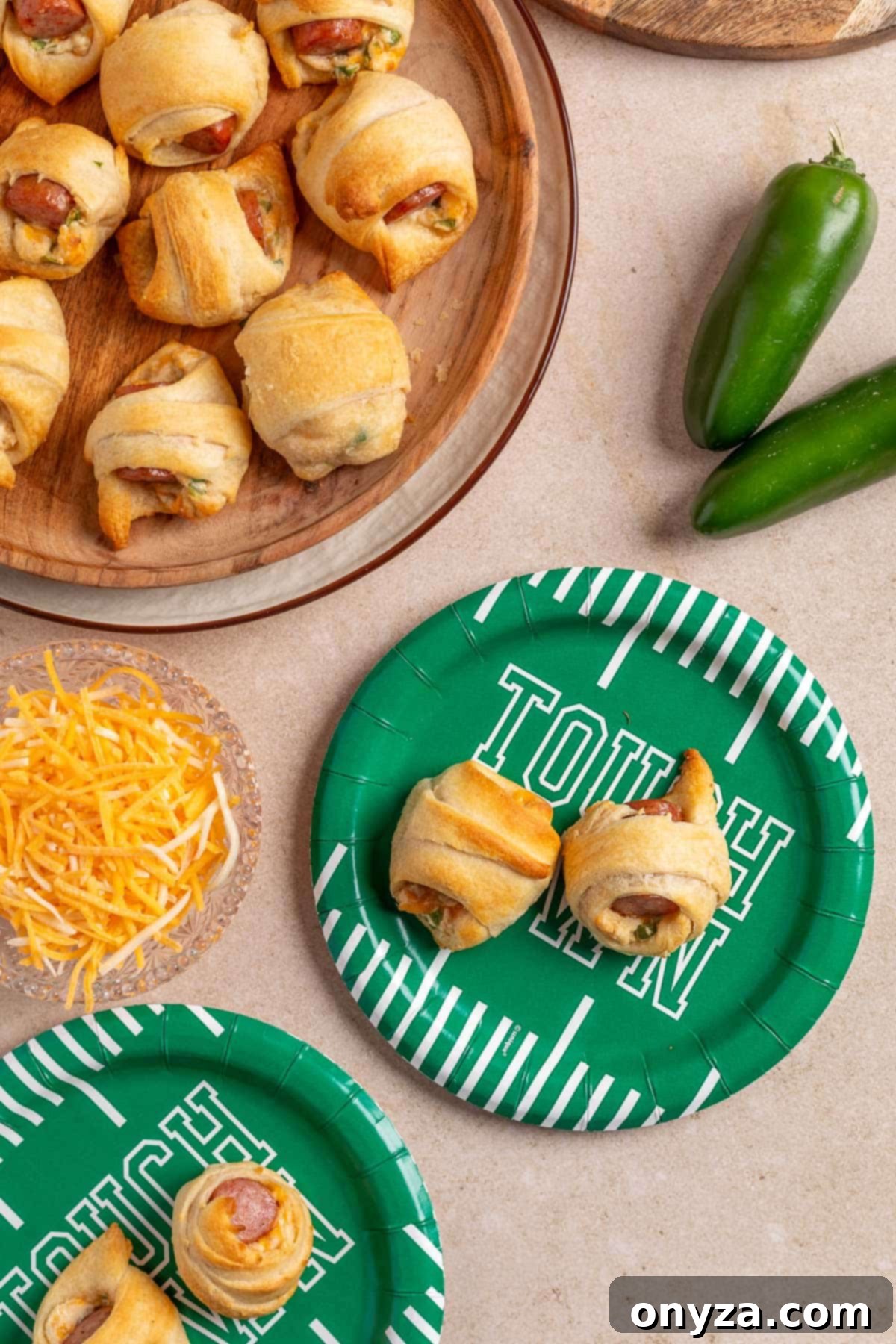 Overhead view of jalapeño popper pigs in a blanket served on a wooden platter and green football-themed paper plates, with shredded cheese and fresh jalapeños nearby on a beige surface.