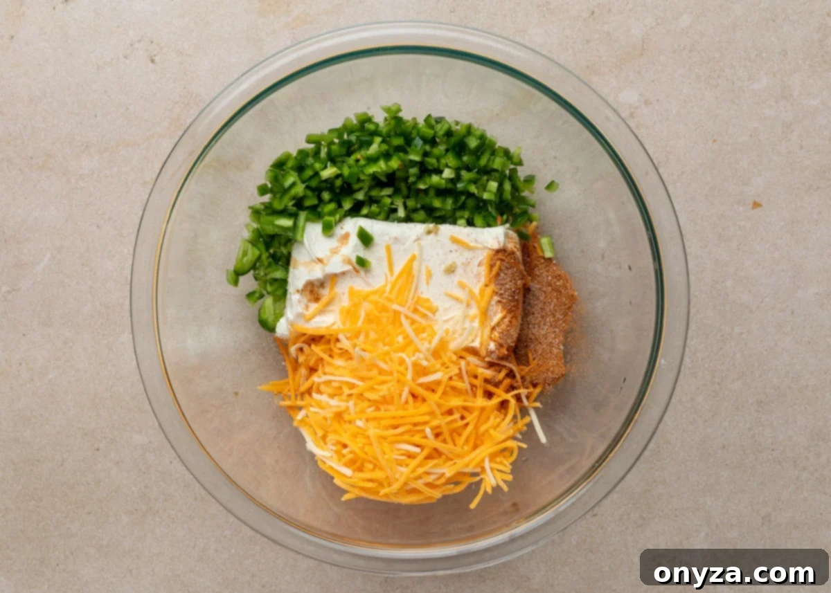 A glass mixing bowl with unblended filling ingredients for Jalapeño Popper Pigs in a Blanket, including diced jalapeños, brick-style cream cheese, shredded cheddar, and Creole seasoning arranged in separate piles.