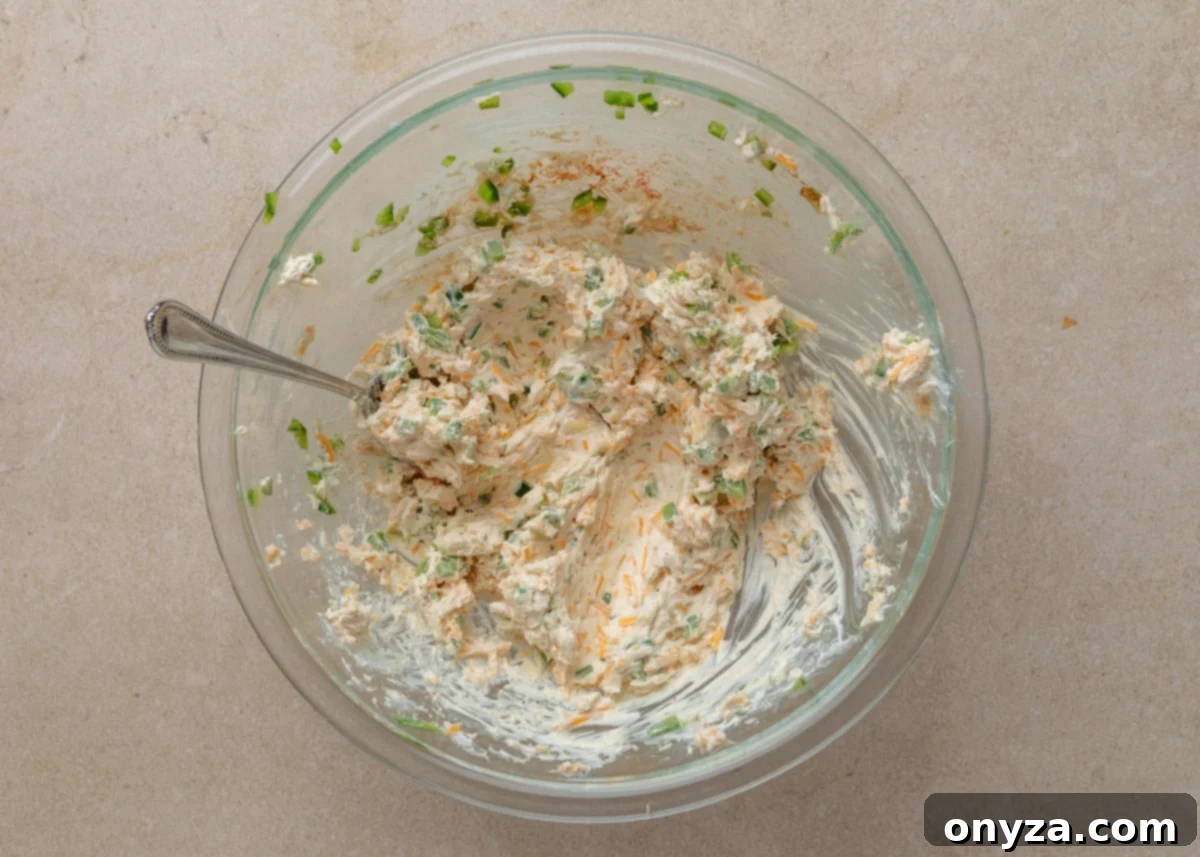 The prepared filling for Jalapeño Popper Pigs in a Blanket in a glass bowl, fully mixed into a creamy, spreadable consistency with visible bits of green jalapeño and orange cheddar.