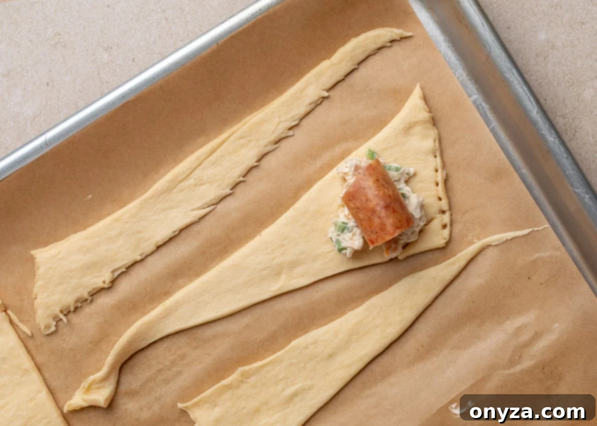 Close-up of crescent roll dough triangle topped with a dollop of jalapeño cream cheese mixture and a piece of smoked sausage, ready to be rolled.