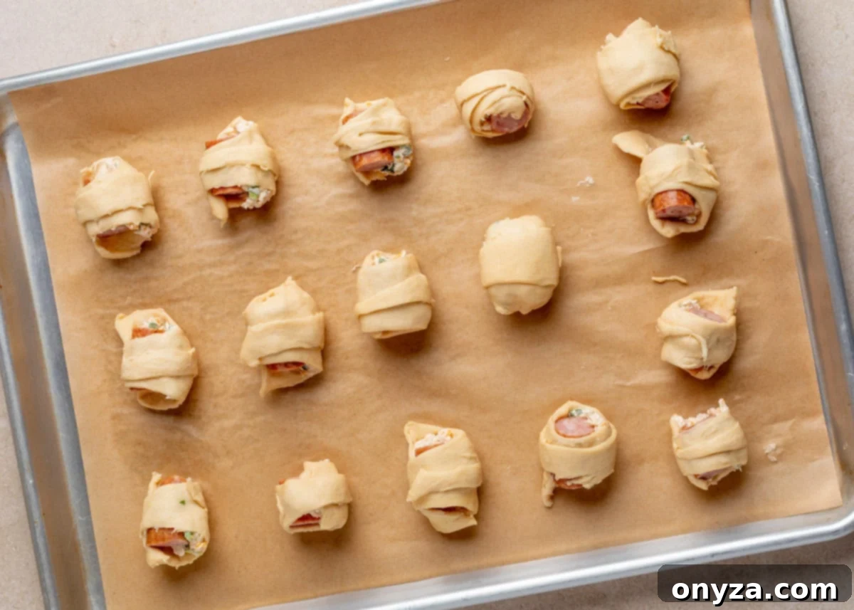 Overhead view of 16 unbaked jalapeño popper pigs in a blanket arranged on a parchment-lined sheet pan, each wrapped in crescent dough and filled with smoked sausage and jalapeño cream cheese mixture.