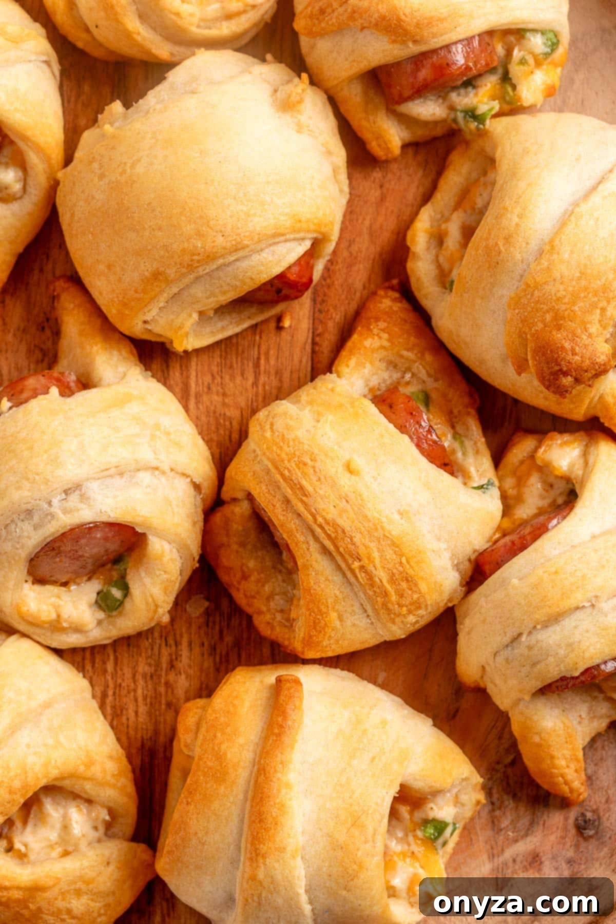 Close-up of freshly baked jalapeño popper pigs in a blanket on a wooden platter, showing golden crescent roll dough wrapped around sausage, melted cheese, and bits of jalapeño.